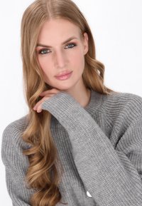 Pulover gri tricotat cu un texturare cu nervuri, guler rotund și mâneci lungi. Părul modelului este ondulat, coafat cu bucle moi și naturale.