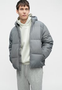 Hallhalli hall puffer-jakk, millel on kapuuts, omab tepitud mustrit, esist zipp ja peene brändimise. Kanda kergelt hoodie ja hallide dressipükste peal.