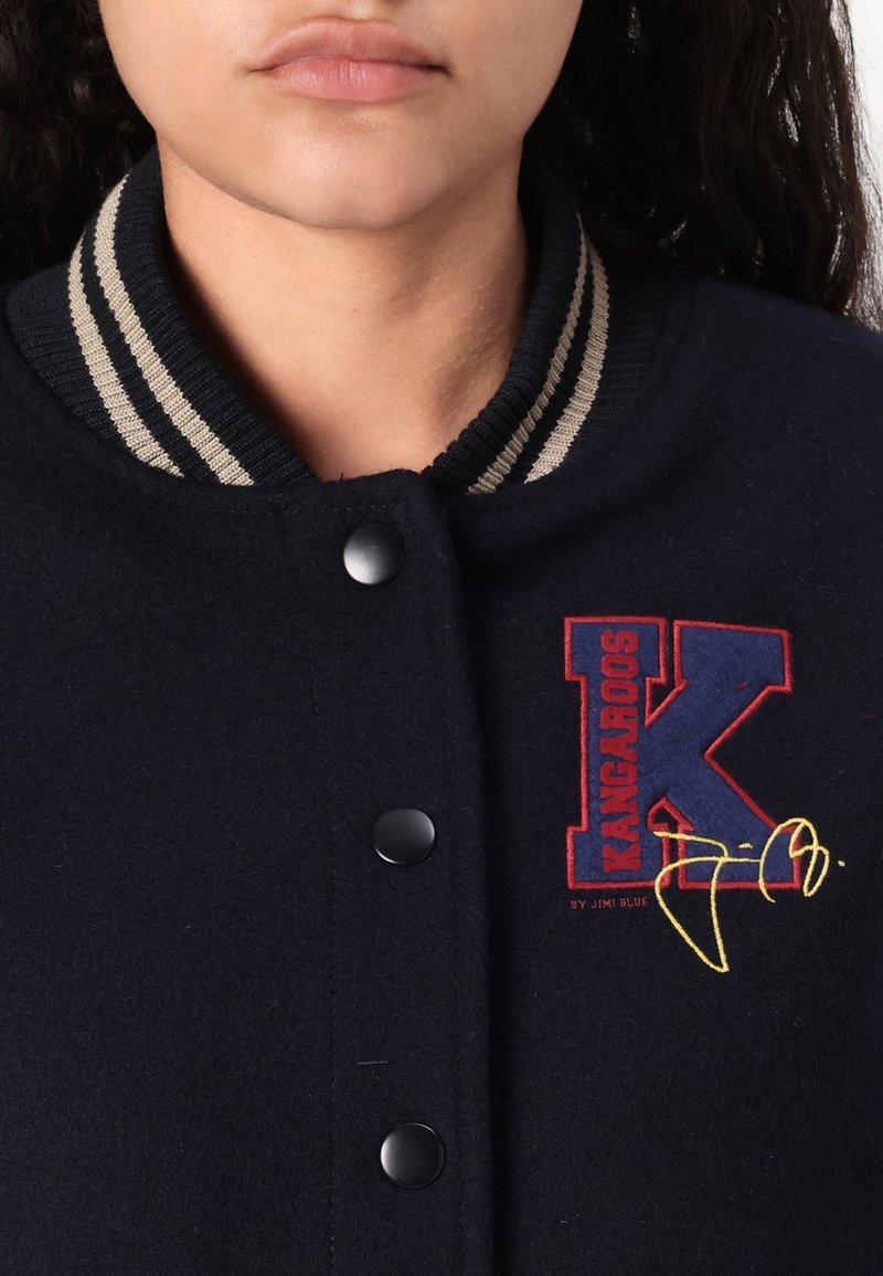 Marineblaue Woll-Collegejacke mit einem gerippten Kragen mit beigen Streifen, schwarzen Druckknöpfen und einem bunten gestickten Logo auf der Brust.