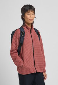 Gequilte koraalfleece jas met hoge kraag, volledige ritssluiting en zijzakken, gedragen met een donkere rugzak.