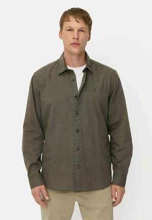Man met olijfgroen overhemd met knopen over een wit T-shirt en beige broek tegen een effen witte achtergrond.