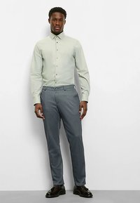 Hellgrünes Hemd mit Knöpfen und Kragen, kombiniert mit grauer Hose und schwarzen Schuhen. Die Passform ist maßgeschneidert und betont eine schlanke Silhouette.