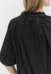 Sort plisseret bluse med et tekstureret stof, rund krave og en særskilt sømdetalje bagpå med en lille metallisk accent.