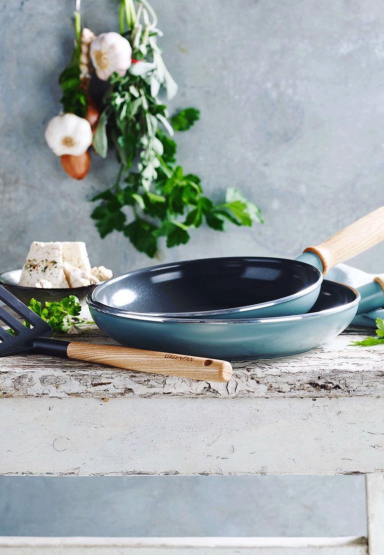 Zwei antihaftbeschichtete Bratpfannen in Türkis und Schwarz mit Holzgriffen, neben einem schwarzen Spatel und frischen Kräutern auf einem rustikalen Tisch.