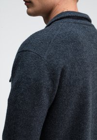 Dunkelgrauer Wollcardigan mit strukturierter Oberfläche, zeigt einen Schalkragen und Details an der Schulternaht, von der Seite betrachtet.