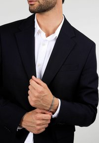 Blazer nero con una camicia bianca sotto, che mette in mostra un bracciale argentato al polso. Il tessuto appare liscio, con un design elegante e su misura.