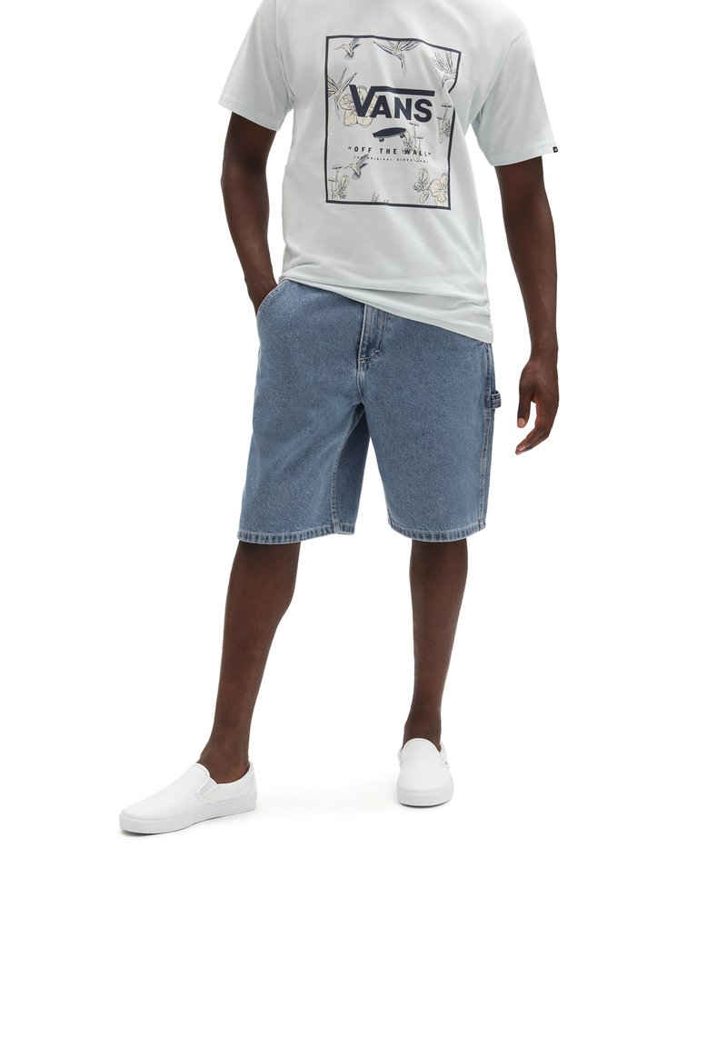 T-shirt grigio chiaro da uomo con logo Vans nero, abbinato a pantaloni corti di jeans blu e scarpe slip-on bianche. I pantaloni corti di jeans presentano tasche laterali.