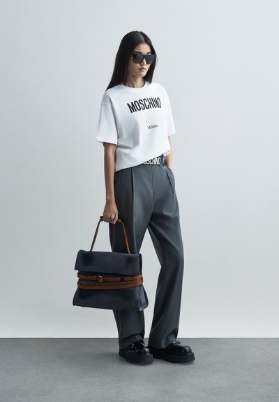 Femeie îmbrăcată cu un tricou alb Moschino, pantaloni gri cu talie înaltă, pantofi cu platformă negri, ochelari de soare negri, ținând o geantă mare gri cu mânere maro.