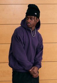 Paarse capuchontrui met een relaxte pasvorm, gestructureerde stof en een zwarte beanie met een wit logo, gecombineerd met een zilveren ketting.