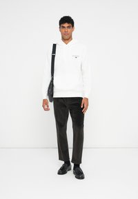 C hoodie branco com um bolso frontal e logótipo, combinado com calças de cotim castanhas e mocassins pretos. O modelo segura uma bolsa preta.