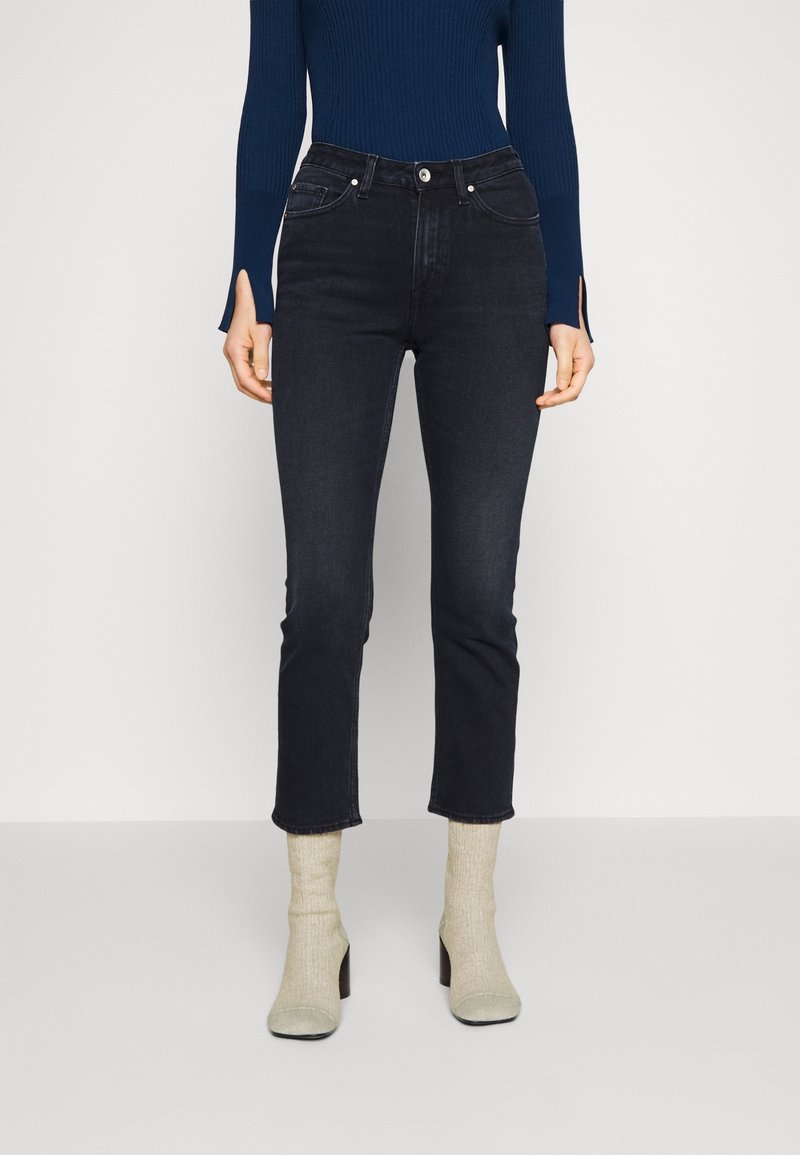Dark denim jeans with a slim fit, zip fly, and five-pocket design. Worn with light-colored, textured ankle boots and a fitted navy top.
