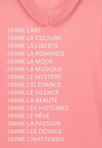 Rózsaszín kapucnis pulóver, fehér szöveges mondatokkal franciául, különböző témák kifejezésére, egyszerű, sans-serif betűtípussal, függőlegesen megjelenítve.