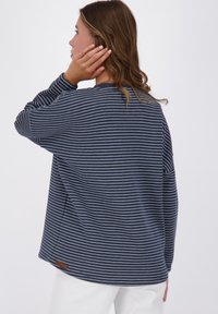 Donna dai capelli mossi che tocca l'orecchio, indossa una maglietta a maniche lunghe blu navy con strisce orizzontali bianche e pantaloni bianchi.