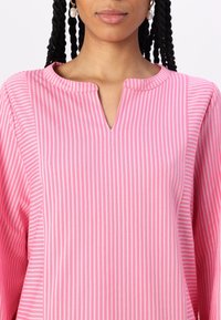 Vaaleanpunainen raidallinen paita, jossa on V-pääntie, ohuet pystysuorat valkoiset raidat ja pitkät hihat. Paita on valmistettu kevyestä kankaasta.