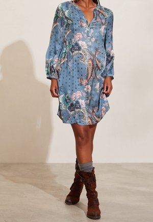 Vrouw draagt een blauwe jurk met bloemen- en paisleymotief, lange mouwen, gecombineerd met grijze sokken en bruine leren laarzen tot halverwege de kuit.