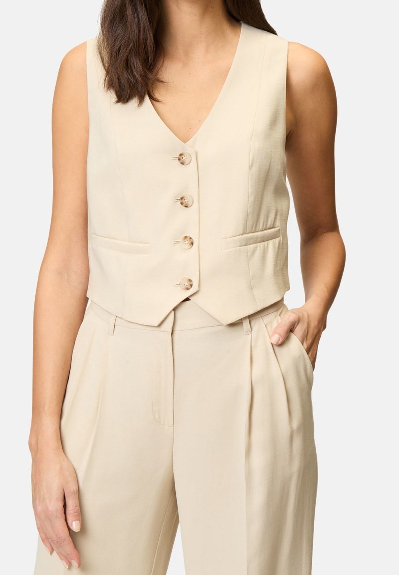 Femme portant un gilet beige sans manches boutonné avec un pantalon plissé taille haute assorti, une main dans la poche, fond neutre.
