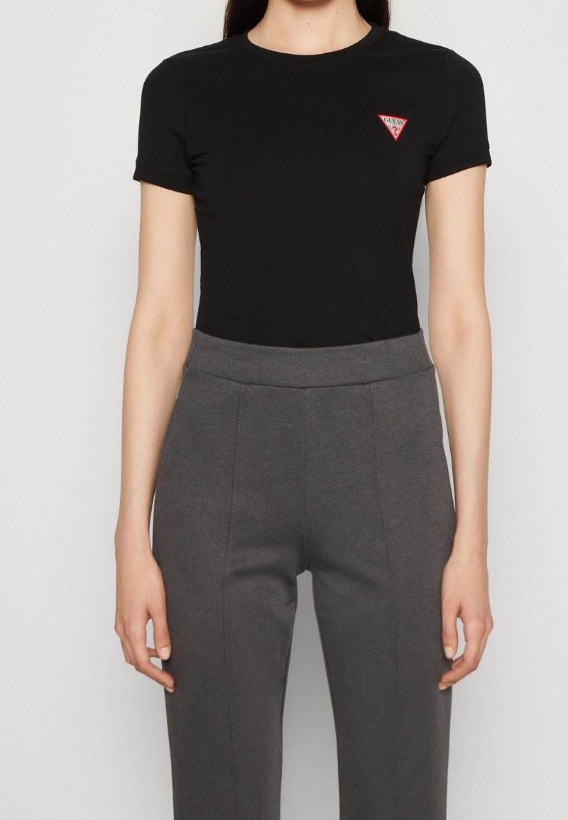 Femme portant un t-shirt noir moulant à manches courtes avec un petit logo triangulaire rouge Guess et un pantalon gris taille haute.