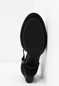 Talons en daim noir avec bout fermé et bride de cheville. Côtés texturés et semelle lisse. Design de talon slim et arrondi. Présenté sur un fond blanc.