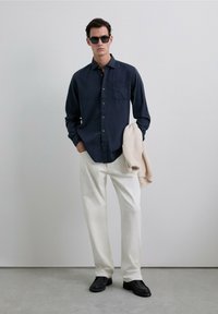 Camisa de botones azul marino, pantalones blancos, zapatos negros y gafas de sol. Suéter de punto suave colgado del brazo. Tejido suave y corte ajustado.