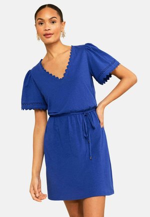 Naine, kes kannab sinist lühikeste varrukatega kleiti lainelise äärise ja vöökohaga lipsuga, seisab ühe käega puusal valge tausta ees.