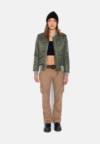 Olivgrüne Satin-Bomberjacke mit grauen Bündchen, schwarzes Crop-Top, braune Cargo-Hosen mit Taschen und schwarze klobige Schuhe. Schwarze Strickmütze.