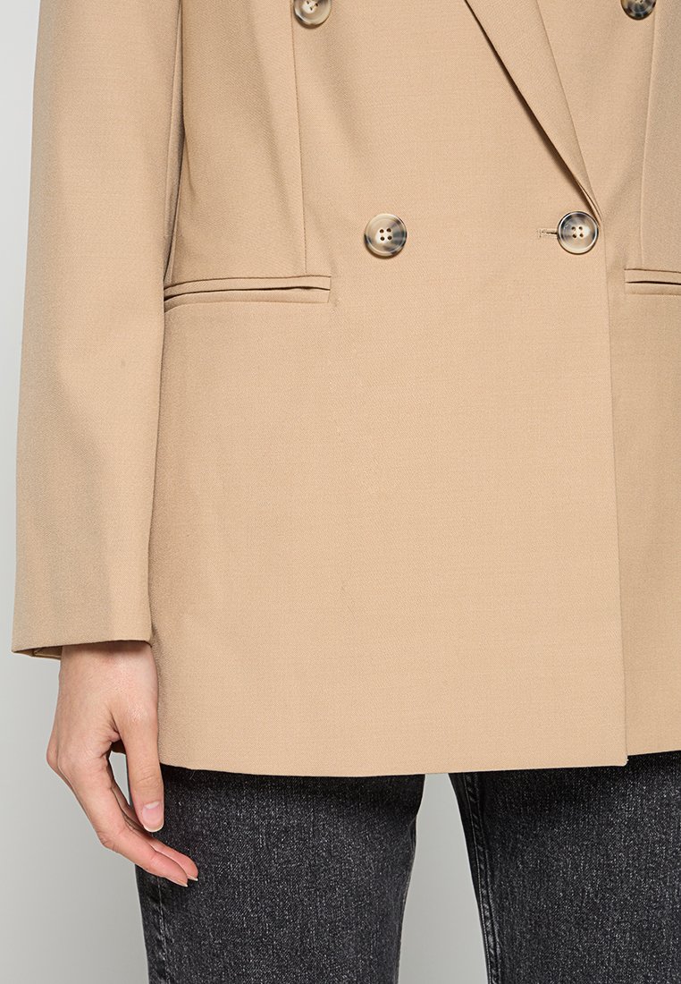 Beige Doppelreiher-Blazer mit zwei großen Knöpfen, Reverskragen und seitlichen Taschen. Der Stoff hat eine glatte Textur und wird mit dunklen Jeans kombiniert.
