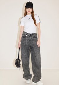 Witte t-shirt met korte mouwen en zwart logo, gecombineerd met oversized zwarte denim jeans. Accessoires omvatten een gestructureerde zwarte handtas en een zwarte pet.