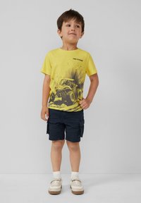 Gelbes T-Shirt mit einem Grafikdesign eines Geländewagens, kombiniert mit dunklen Cargo-Shorts. Weiße Schuhe mit beigen Akzenten und strukturierte Socken.