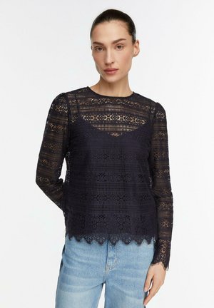 Kobieta z uczesanymi włosami, ubrana w czarną koronkową bluzkę z długim rękawem na czarnym topie na ramiączkach i jasnoniebieskie dżinsy, stojąca na białym tle.