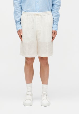 Homme portant un short crème avec cordon de serrage, une chemise à manches longues bleu clair boutonnée, des chaussettes blanches et des baskets blanches, se tenant devant un arrière-plan uni.
