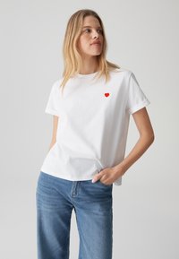 T-shirt bianco in cotone con un piccolo ricamo a forma di cuore rosso sulla parte sinistra del petto, maniche arrotolate e vestibilità rilassata. Abbinato a jeans blu.