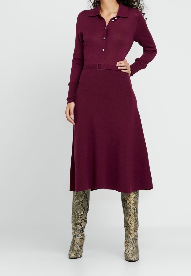 Robe en maille bordeaux à manches longues, col, détail à boutons et taille ceinturée. Portée avec des bottes montantes à imprimé serpent.