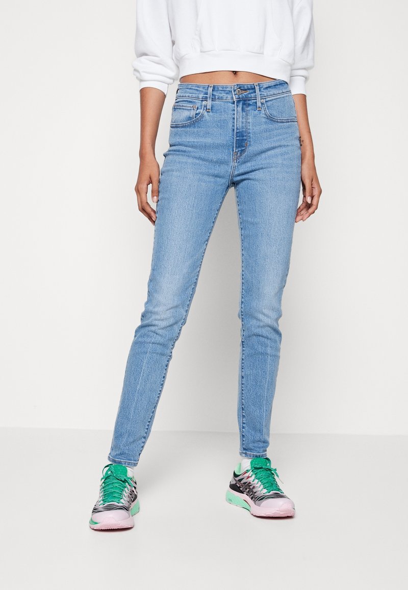 Personne portant un jean skinny bleu clair, un sweat court blanc et des baskets multicolores avec des lacets verts debout sur un sol clair uni.