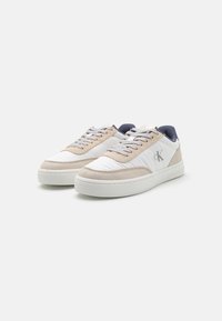 Lightweight sneaker with white synthetic upper, beige suede accents, gray laces, and a rubber sole. Features a logo on the side and a textured back.