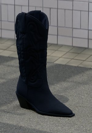 Fekete cowboy csizma díszítő varrásokkal és dőlt sarokkal, szürke járdán, csempézett fal közelében.