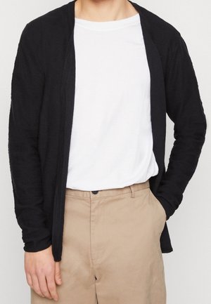 Personne portant une chemise blanche rentrée dans un pantalon beige avec une main dans la poche et un cardigan noir ouvert.