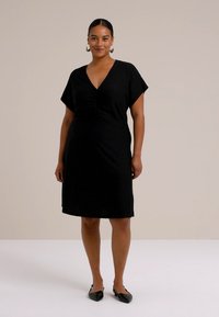 Leende kvinna som bär en svart klänning med korta ärmar, står mot en ljus beige bakgrund och visar en avslappnad passform och elegant stil.
