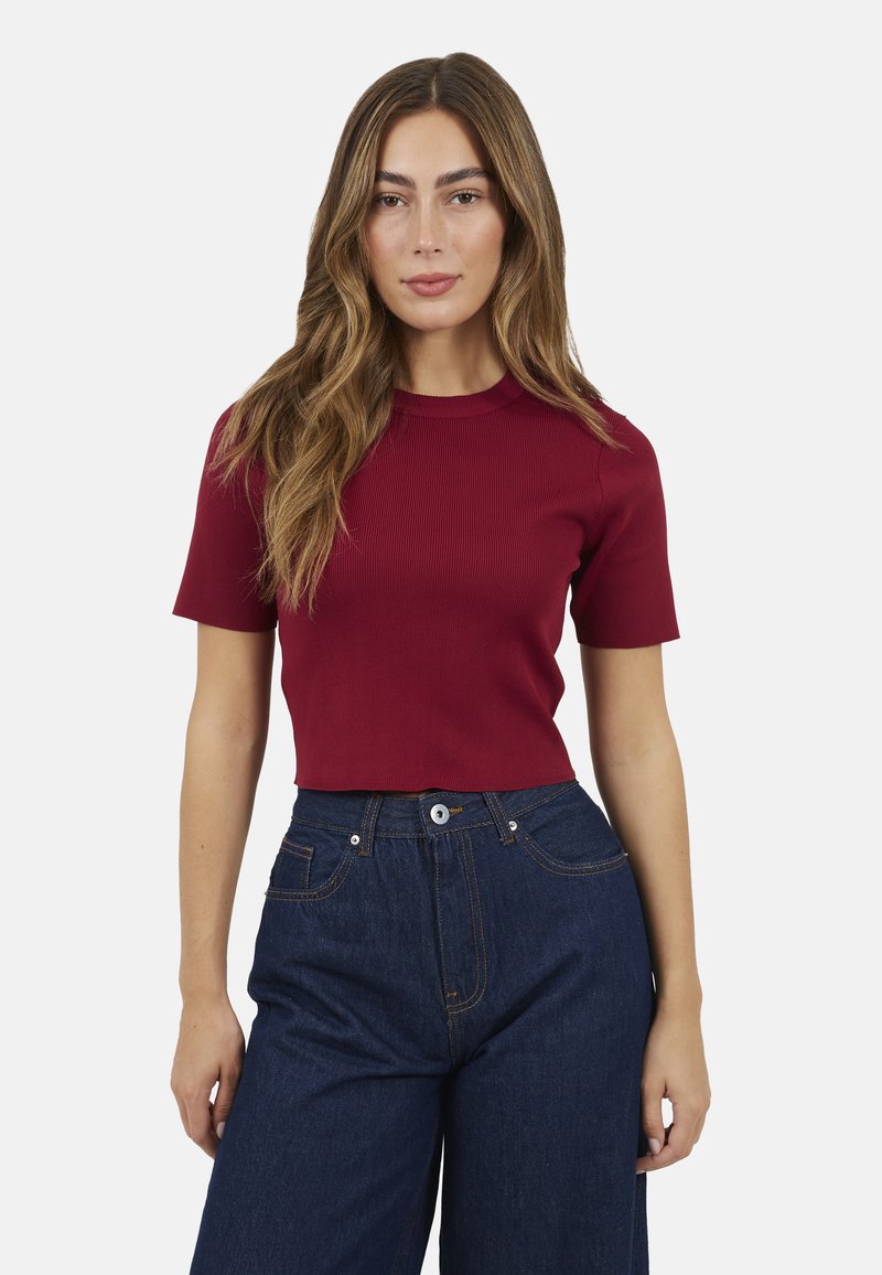 Vrouw met lang golvend haar, gekleed in een bordeauxrood crop top met korte mouwen en een donkerblauwe high-waisted jeans, tegen een witte achtergrond.
