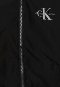 Giacca a vento nera con zip a tutta lunghezza, con un discreto logo Calvin Klein Jeans bianco nella zona del petto. Texture liscia.