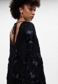 Melnā kleita ar ziedu izšuvumiem, zemu aizmuguri un aukliņas aizdari. Grezna tekstūra un plūstošas piedurknes izceļ sarežģīto dizainu.