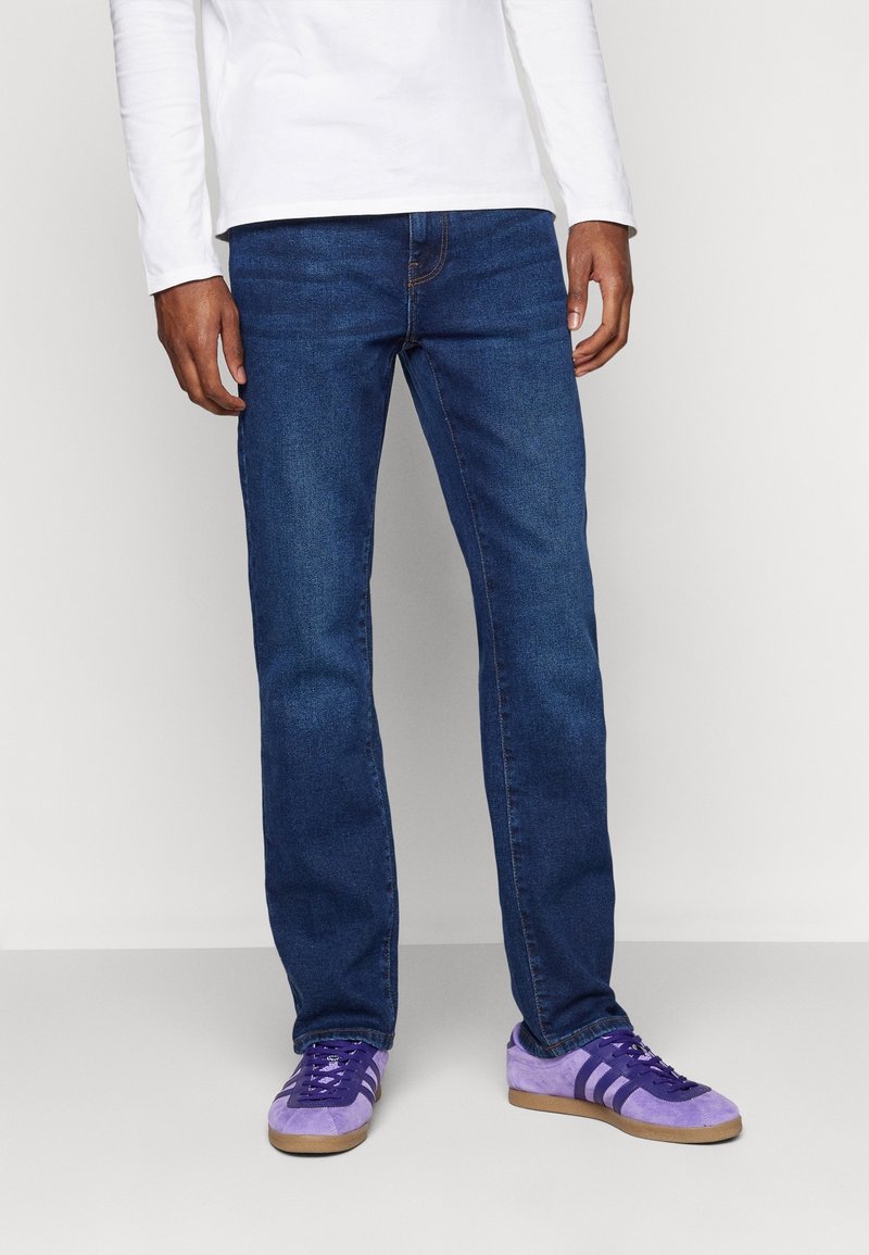Homme portant un jean bleu foncé, une chemise blanche à manches longues et des baskets violettes avec des rayures blanches, debout devant un fond uni.