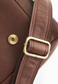 Correa de bolsa de cuero marrón con accesorios de latón. Presenta una superficie texturizada, costuras en contraste y un detalle de botón a presión redondo.