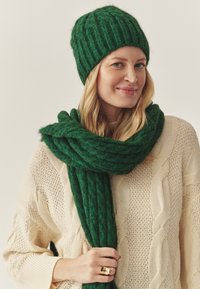 Grøn strikket hue og matchende tørklæde lavet af tekstureret materiale, båret over en krèmefarvet kabelstrikket sweater. Guld ring synlig på hånden.