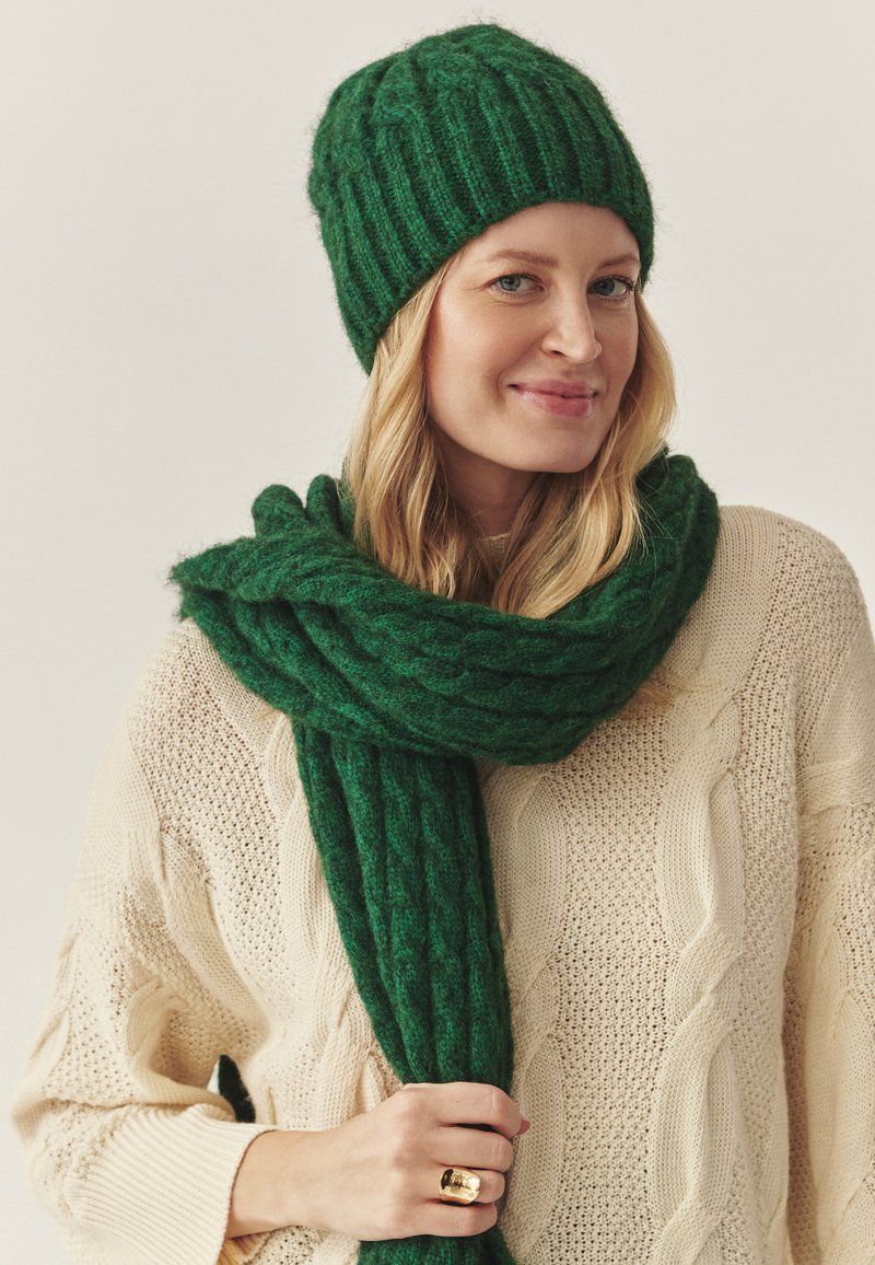Grøn strikket hue og matchende tørklæde lavet af tekstureret materiale, båret over en krèmefarvet kabelstrikket sweater. Guld ring synlig på hånden.