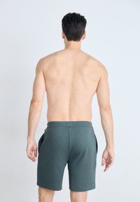 En mann med kort, mørkt hår står uten skjorte, vender bort, iført mørkegrønne shorts mot en lys, ensfarget bakgrunn.