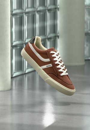 Barna-fehér alacsony szárú sneaker fehér fűzőkkel és "BOSS" logóval, modern üvegtéglafal és polírozott padló előtt kiállítva.