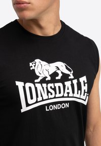 Juodas be rankovių marškinėliai iš medvilnės audinio, išsiskiriantys ryškiu baltu liūto logotipu ir užrašu "LONSDALE LONDON", atspausdintu priekyje.