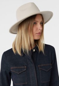 Cappello in feltro di lana con tesa larga color beige chiaro, caratterizzato da una texture liscia e un design strutturato. Giacca di jeans con cuciture a contrasto.