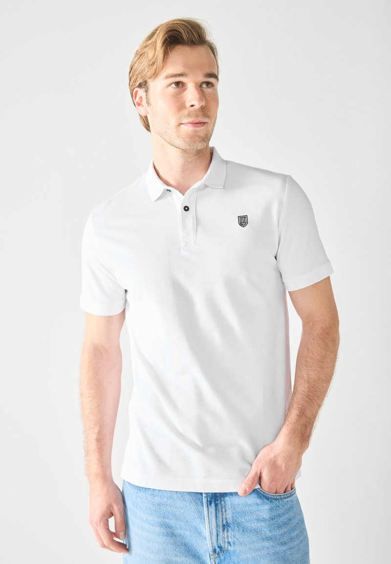 Chemise polo blanche avec un col, deux boutons noirs, et un logo de blason brodé sur la poitrine gauche. Portée avec un jean en denim bleu clair.