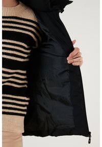 Black puffer jacket with a smooth inner lining, displayed open. A striped knit sweater in beige and black is worn underneath.
